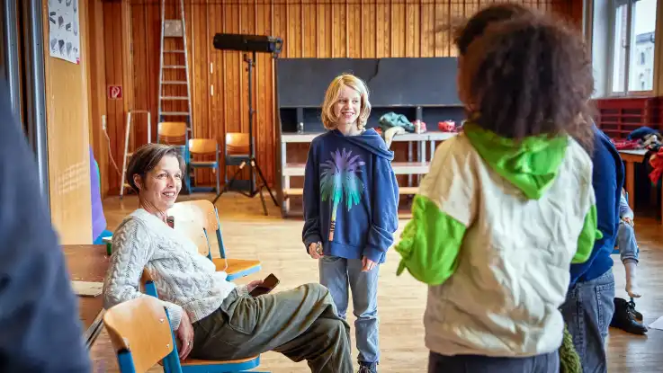 In einem holzvertäfelten Raum mit hoher Decke sitzt eine Frau lächelnd auf einem Stuhl, um sie herum haben sich einige Kinder versammelt.