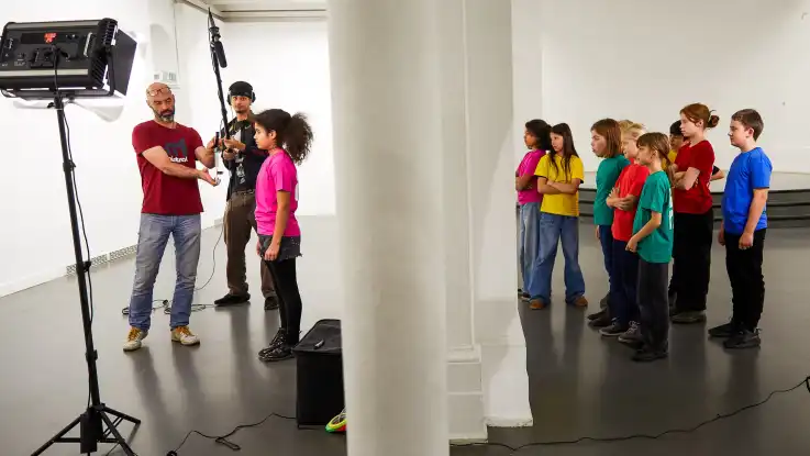 Eines der Kinder, ein Mädchen in pinkem T-Shirt, wartet auf ihren Einsatz. Neben ihr steht ein junger Mann mit einem Richtmikrophon.