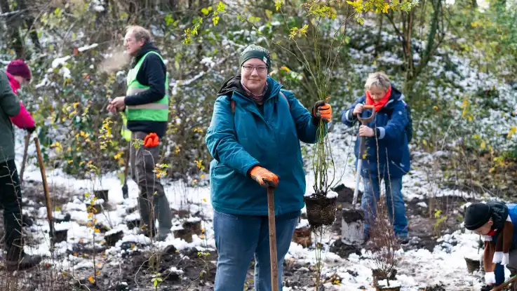 Eine Gruppe hebt mit Sparten kleine Erdlöcher im Waldboden aus – eine der Frauen hält stolz einen Setzling in der Hand. 