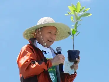  Ein älterer Mann mit Strohhut hält in der einen Hand ein Mikrophon und in der anderen einen Setzling. 