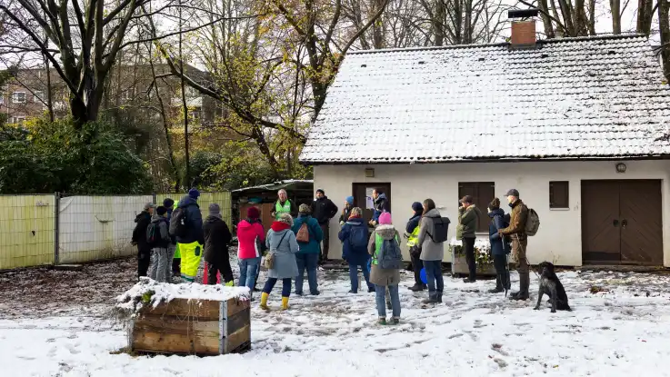 Vor einem kleinen Häuschen mit Satteldach hat sich eine größere Gruppe um einen Mann mit grüner Warnweste geschart. 