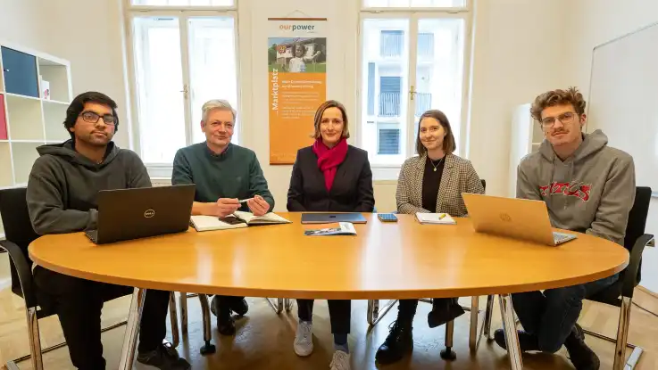Gruppenfoto: Fünf Personen unterschiedlichen Alters sitzen an einem großen, runden Holztisch und blicken in Richtung Kamera. 