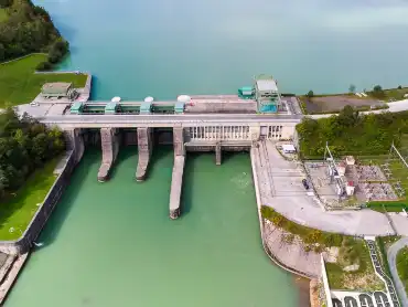 Luftaufnahme: An einem Stausee steht eine Staumauer mit drei Überläufen und einem kleinen Umspannwerk.