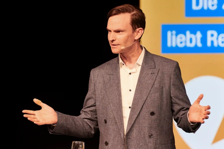 Andreas Wieg steht mit ausgebreiteten Armen an einem Podium und spricht.