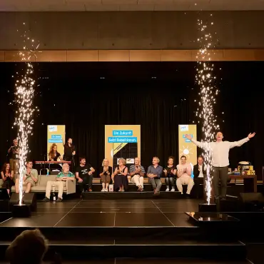 Auf einer Bühne sitzen im Hintergrund zahlreiche Menschen, ein Mann im Vordergrund hebt die Arme, während ein kleines Bühnenfeuerwerk die Szene illuminiert.