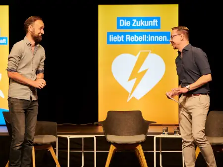 Zwei jüngere Männer stehen auf einer Bühne und lachen sich freundlich an.