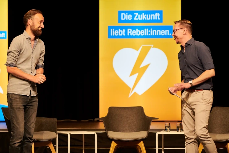 Zwei jüngere Männer stehen auf einer Bühne und lachen sich freundlich an.