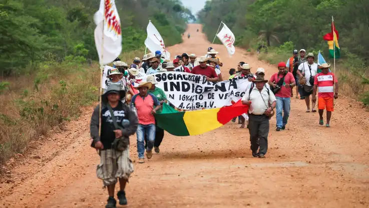 Eine Gruppe von Demonstranten mit Fahnen und Bannern marschiert über eine rote Staubpiste, die sich durch den Wald zieht. 