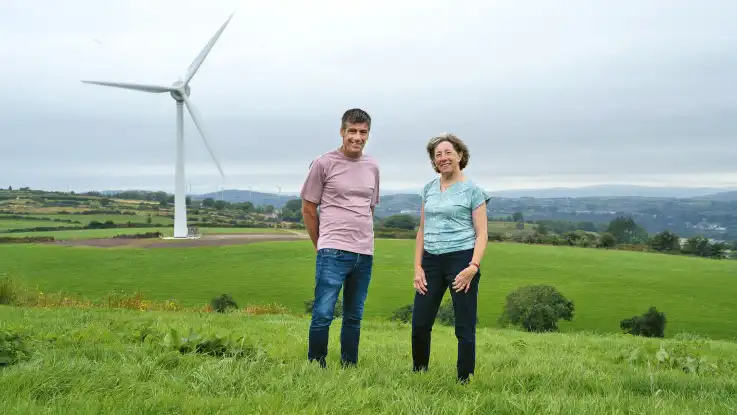 Der Mann posiert nun mit einer Frau in blauem Oberteil vor einer Windkraftanlage, beide lächeln stolz in die Kamera. 