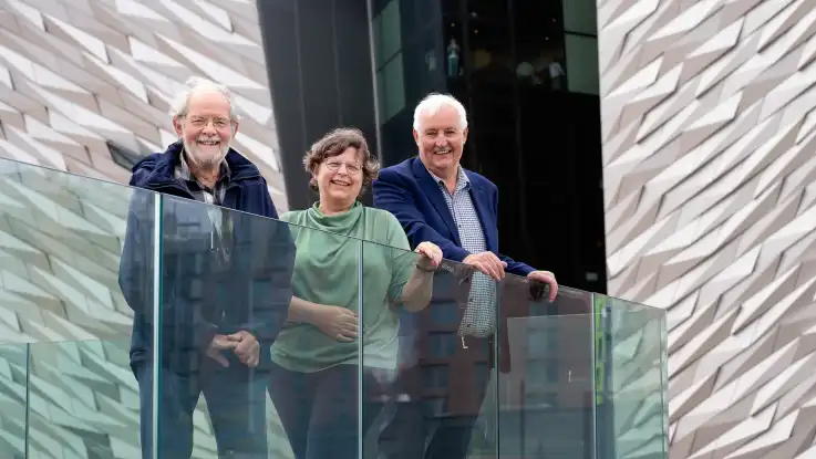 Eine Frau und zwei Männer lehnen lächelnd an einer Glasbrüstung. Den Hintergrund bildet eine moderne Hausfassade mit polygonaler Struktur.