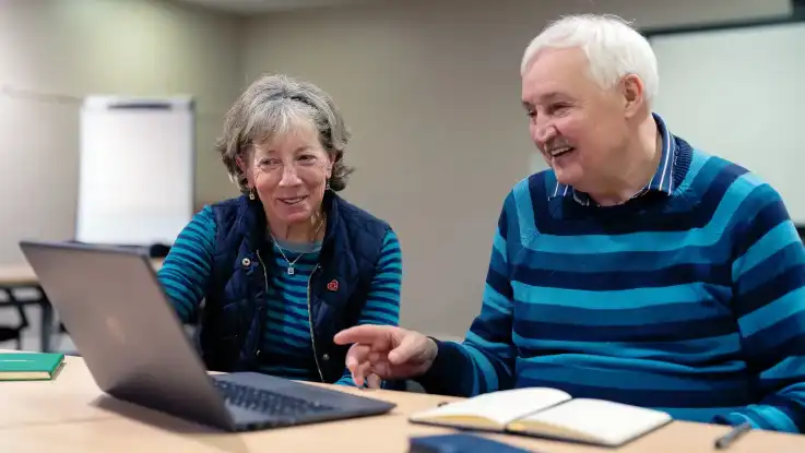 Zwei ältere Personen, ein Mann und eine Frau, sitzen an einem Laptop. Beide tragen blau gestreifte Kleidung. 