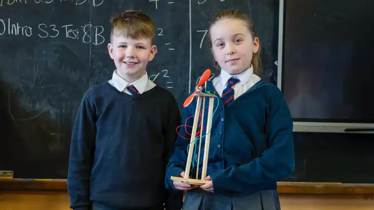 Ein Junge und ein Mädchen stehen vor einer Tafel, das Mädchen hält stolz eines der Windräder als wäre es eine Trophäe.