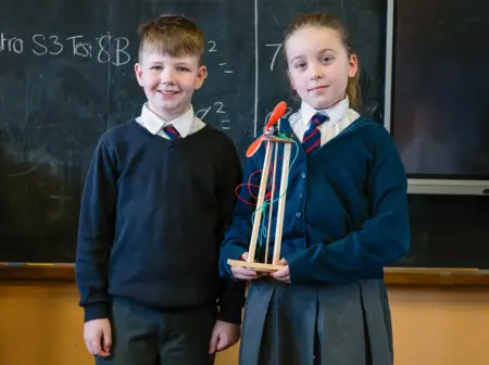 Ein Junge und ein Mädchen stehen vor einer Tafel, das Mädchen hält stolz eines der Windräder als wäre es eine Trophäe.