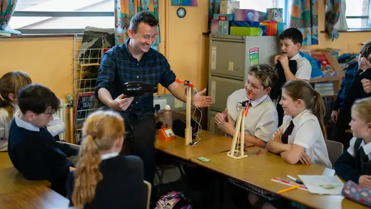 Der Mann im Karo-Hemd zielt mit einem Fön auf eines der Windrad-Modelle, um ihn herum haben sich staunende Kinder versammelt. 
