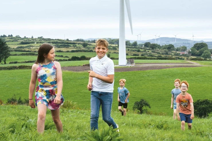 Five children are playfully running up a green hill; wind turbines stand in the landscape behind them. 