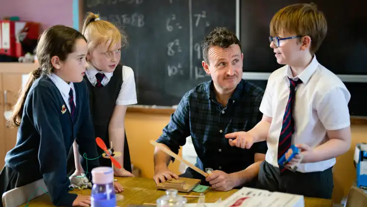 Der Mann sitzt nun mit Schüler:innen an einem der Tische: zwei Mädchen fixieren einen Jungen, der mit einer Säge hantiert, mit ernstem Blick. 