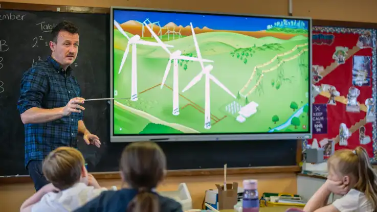 Ein Mann im blau-schwarzen Karo-Hemd steht in einem Klassenzimmer, das Smartboard neben ihm zeigt die Illustration einer Windkraftanlage. 