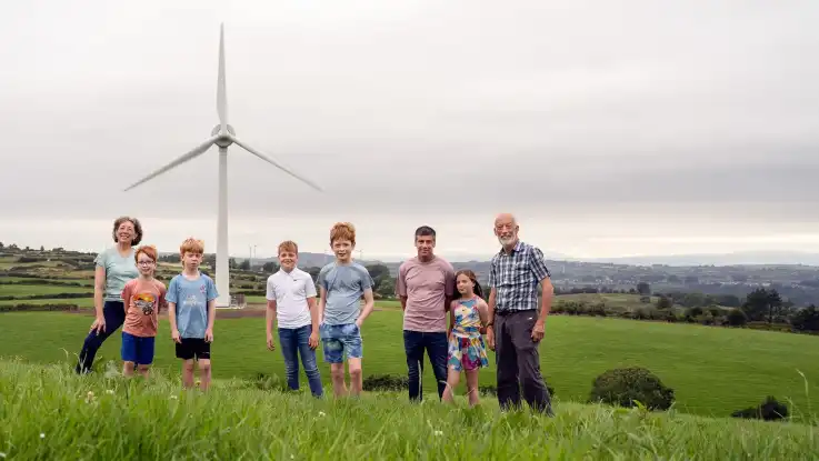 Eine Gruppe von fünf Kindern und drei Erwachsenen posiert vor einer Windkraftanlage. 