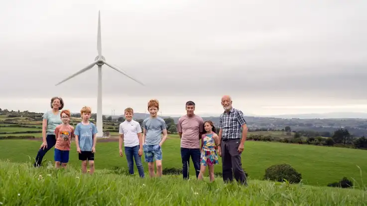 Eine Gruppe von fünf Kindern und drei Erwachsenen posiert vor einer Windkraftanlage. 