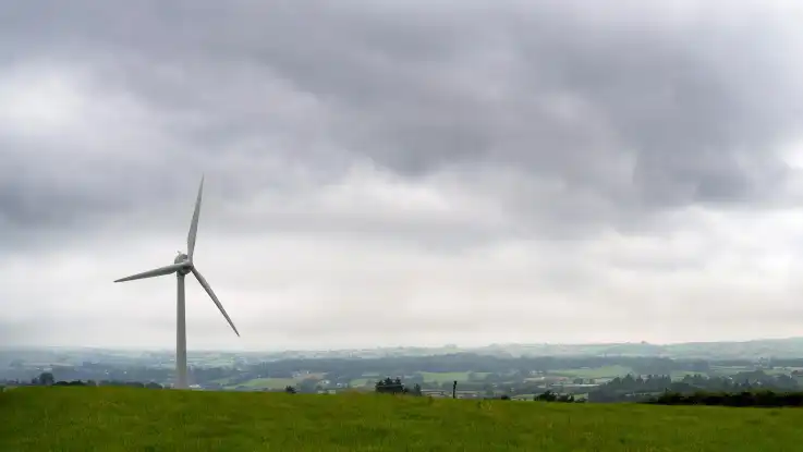 Dunkelgraue Wolken hängen über einer grünen, hügeligen Landschaft – links im Bild steht ein einzelnes Windrad. 