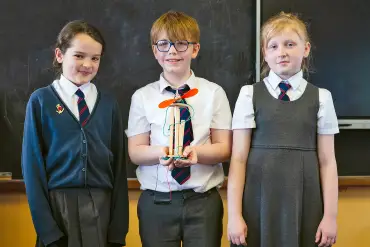 Drei Kinder in Schuluniform posieren stolz mit dem Modell einer Windkraftanlage. 