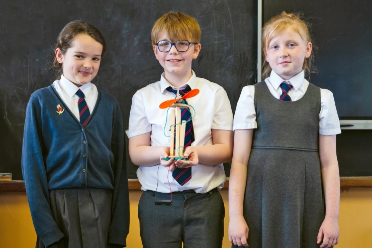 Drei Kinder in Schuluniform posieren stolz mit dem Modell einer Windkraftanlage.