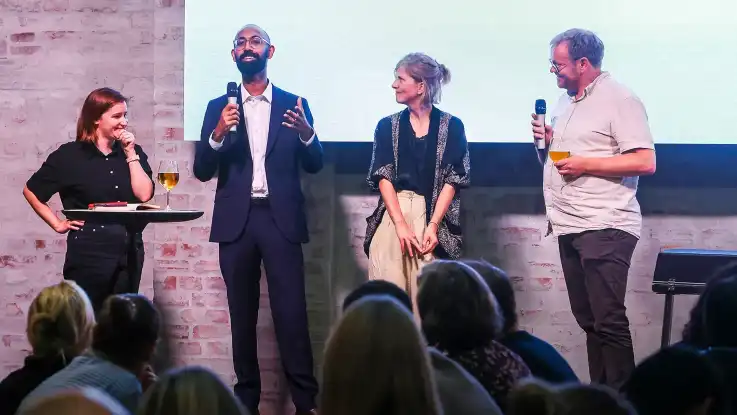 Eine der Personen auf der Bühne, ein Mann mit Glatze und schwarzem Vollbart, spricht zum Publikum. 