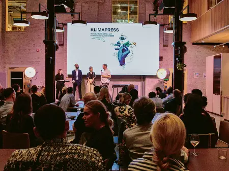 In einer kleinen Halle stehen vier Personen auf der Bühne, davor sitzt das Publikum locker verteilt an Tischen. 