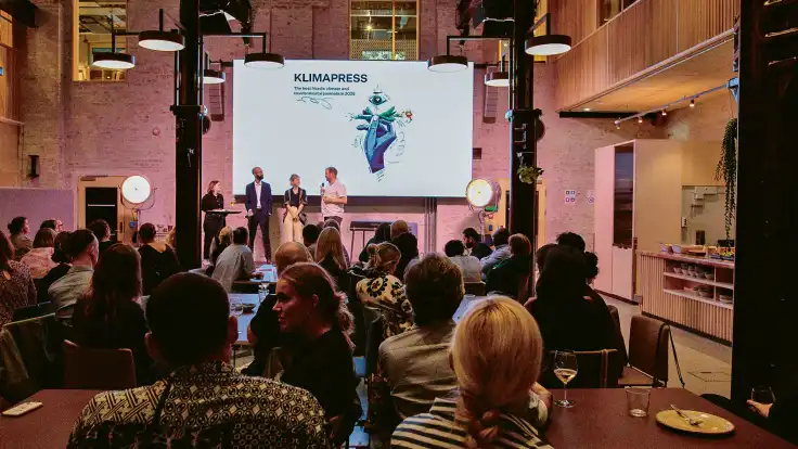 In einer kleinen Halle stehen vier Personen auf der Bühne, davor sitzt das Publikum locker verteilt an Tischen. 