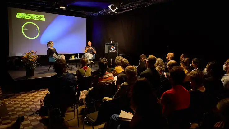 Auf einer Bühne sitzt vor vollbesetztem Saal eine Frau und ein Mann im Gespräch.