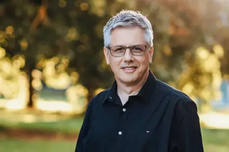 Porträt: Ein Mann mit grauem Haar und Brille steht im Freien und lächelt freundlich.