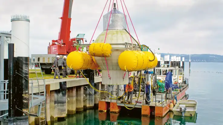Eine kugelförmige Betonkonstruktion wird von Monteuren mithilfe eines Autokrans im türkisfarbenen Wasser eines Sees versenkt.