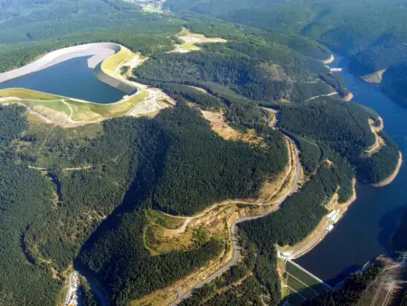 Luftaufnahme: Auf einem Berg befindet sich ein riesiges Wasserbecken mit betonierten Wänden. Im Tal darunter erstreckt sich ein Stausee. 