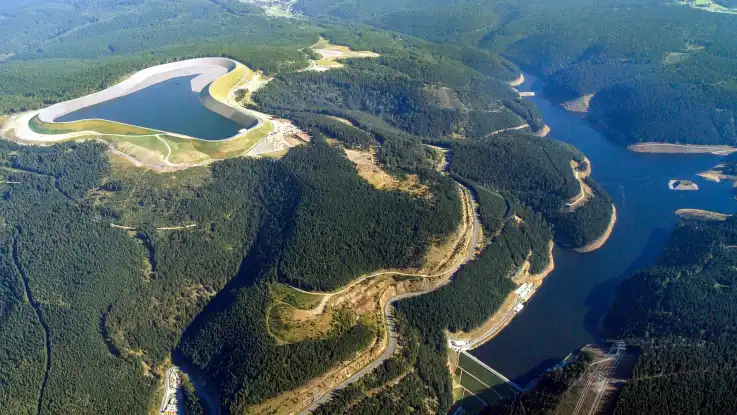 Luftaufnahme: Auf einem Berg befindet sich ein riesiges Wasserbecken mit betonierten Wänden. Im Tal darunter erstreckt sich ein Stausee. 