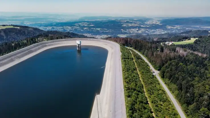 Auf einem Plateau befindet sich ein ovales Betonbecken. Aus dem Wasser ragt ein riesiger Überlauf. 