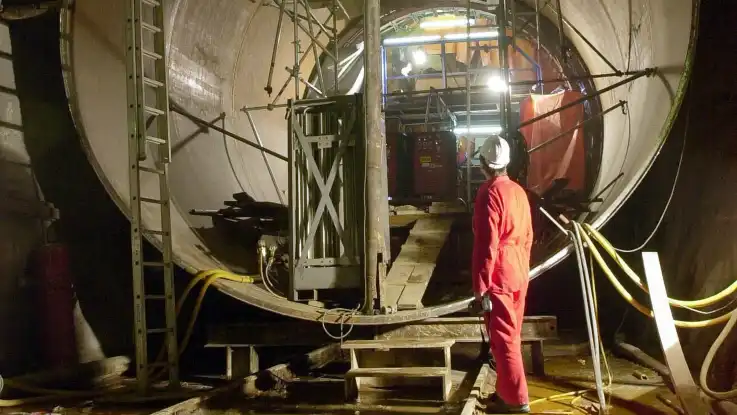 Ein Mann in rotem Overall und mit Schutzhelm auf dem Kopf blickt in eine überdimensionierte Röhre. 