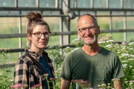 Eine junge Frau mit hochgestecktem Zopf steht neben einem älteren Mann – beide tragen eine ähnliche Brille und lächeln freundlich. 