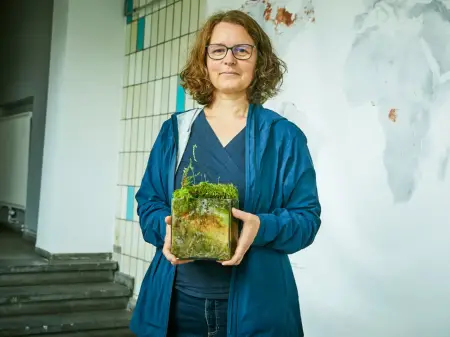 Eine junge Frau steht vor einer Weltkarte und hält einen Glaskasten mit einem kleinen Biotop darin in der Hand. 