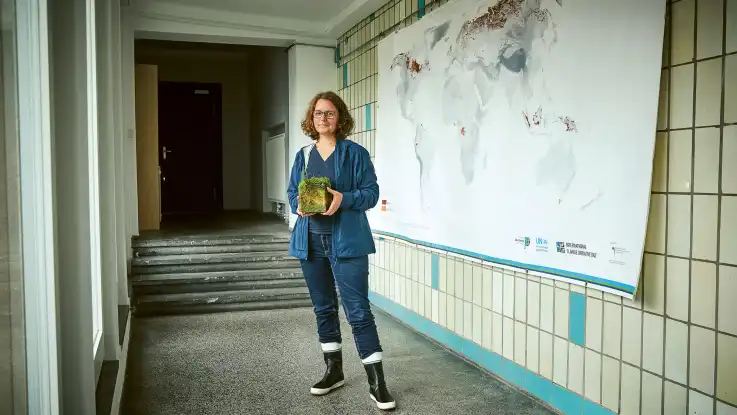 Eine Frau mittleren Alters steht mit Gummistiefeln in einem gekachelten Raum vor einer Wandkarte. 