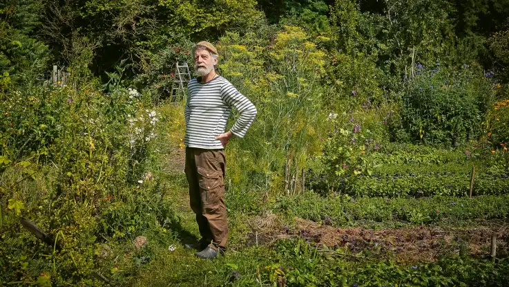 Ein älterer Mann mit grauem Vollbart, Strickmütze und gestreiftem Oberteil steht in einem kleinteiligen Beet. 