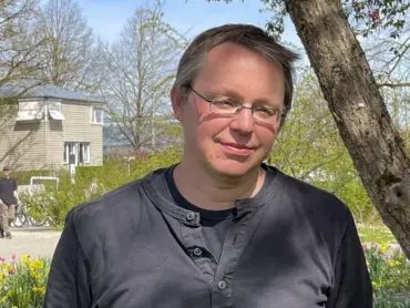 Ein Mann mittleren Alters mit grau meliertem Haar und Brille steht im Freien und lächelt freundlich. 