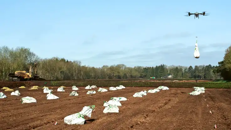 Eine Drohne schwebt mit einem weißen Plastiksack über einem Torf-Feld, auf dem bereits zahlreiche Säcke verteilt liegen. 
