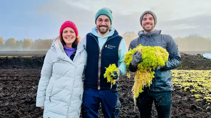 Eine Frau und zwei Männer in den 30ern posieren lachend auf einem Torf-Feld. Die Männer haben Torfbüschel in der Hand. 