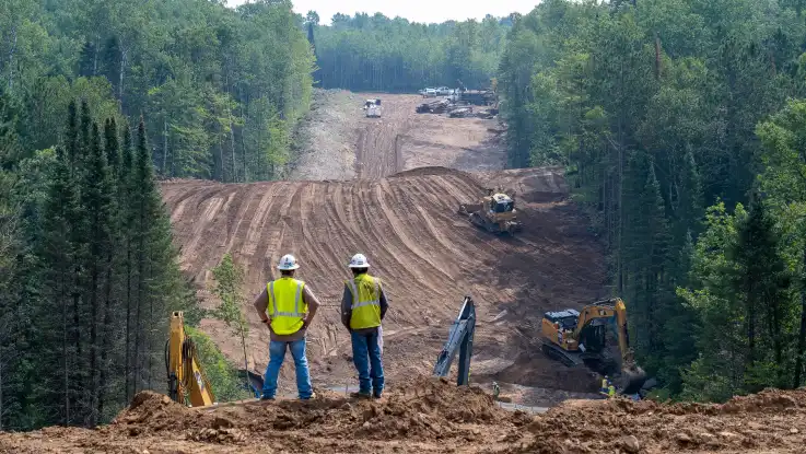 Bauarbeiter schauen auf eine breite, von Baggern gerodete Schneise, die durch einen dichten Wald verläuft.