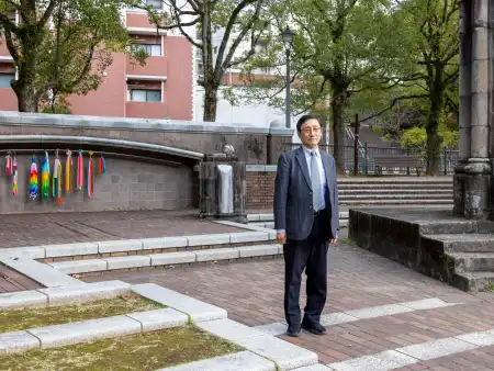 Ein Mann steht neben einer Ruine, hinter ihm hängen bunte Kranich-Girlanden vor einer Steinwand. 