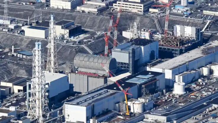Schräg von oben: zwischen großen rechteckigen Reaktorhallen und Abluftkaminen stehen zahlreiche rot-weiße Kräne.