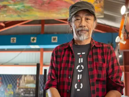 Ein Mann mit grauem Schnurr- und Kinnbart schaut empathisch zur Kamera, hinter ihm eröffnet sich eine kleine Halle in der farbenfrohen Bilder hängen. 