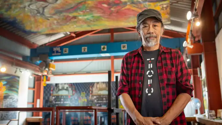 Ein Mann mit grauem Schnurr- und Kinnbart schaut empathisch zur Kamera, hinter ihm eröffnet sich eine kleine Halle in der farbenfrohen Bilder hängen. 