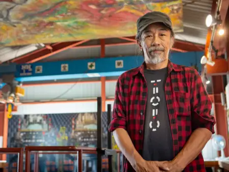 Ein Mann mit grauem Schnurr- und Kinnbart schaut empathisch zur Kamera, hinter ihm eröffnet sich eine kleine Halle in der farbenfrohen Bilder hängen. 