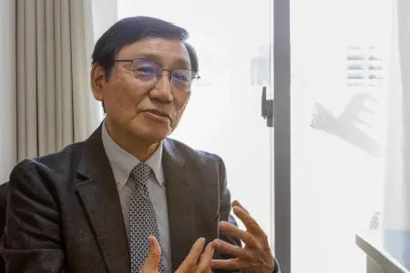 Ein japanischer Mann im Anzug mit Brille und Seitenscheitel erzählt gestenreich seine Geschichte. 